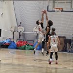 Gracz UJK o nazwisku Flisiak, biegnie w kierunku kosza drużyny AZS AGH Kraków i wymierza rzut, podczas gdy zawodnik drużyny przeciwnej usiłuje przeszkodzić w zdobyciu punktu